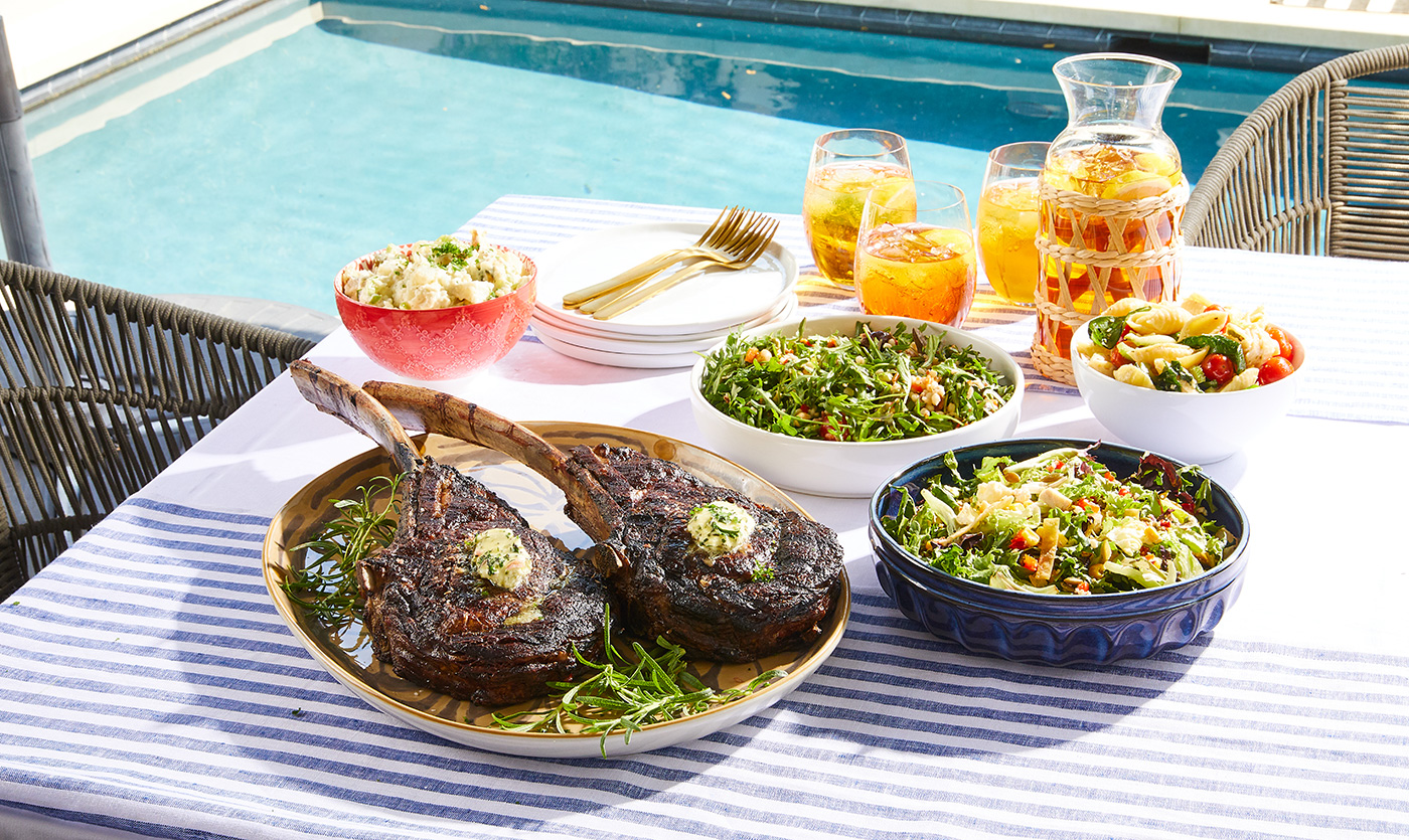 Tomahawk Steaks with sides and drinks on a table by the pool
